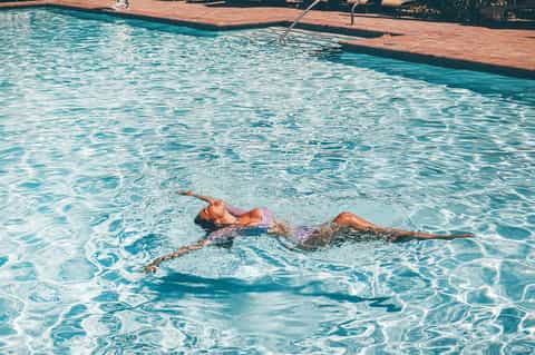 Woman floating peacefully in crystal-clear turquoise pool water with wooden dock and green landscape