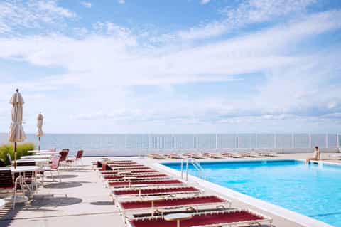 Outdoor pool deck with burgundy lounge chairs, umbrellas, and ocean view behind white fence