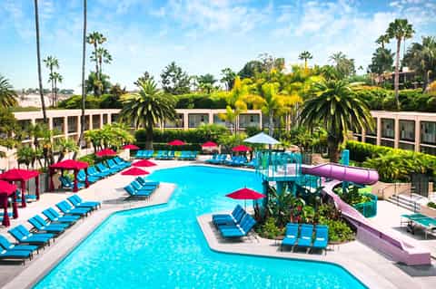 Resort pool complex with turquoise water, blue lounge chairs, red umbrellas, and palm trees under clear sky