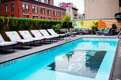 Urban rooftop pool with white lounge chairs, colorful striped cabanas, and brick buildings backdrop