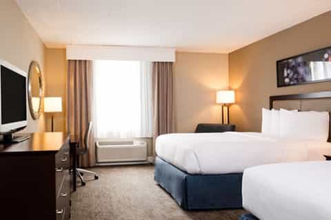 Hotel room with two queen beds, warm neutral tones, work desk, TV, and window with curtains.