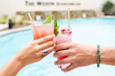 Two colorful cocktails with mint garnish held by hands at poolside during daytime
