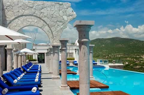 Luxurious poolside terrace with blue loungers, stone columns, arches, and mountain views under clear sky