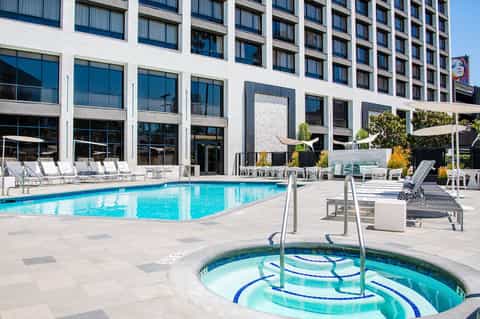 Outdoor pool deck with lounge chairs, hot tub in foreground, and modern hotel building facade