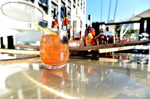 Cocktail with orange garnish on outdoor bar counter with bottles and staff in background