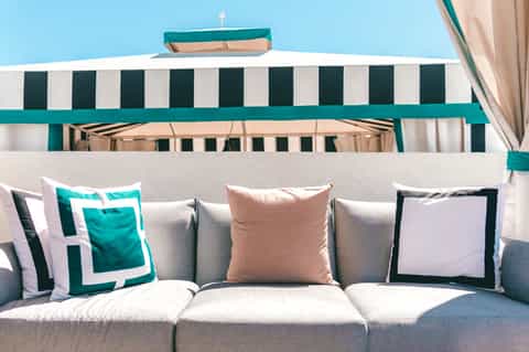Beachside lounge sofa with teal and white striped awning and decorative pillows against blue sky