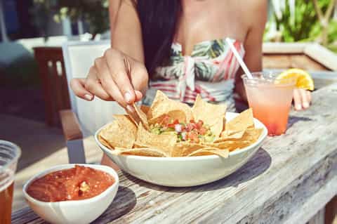 Person reaching for tortilla chips with salsa and tropical drink at poolside table