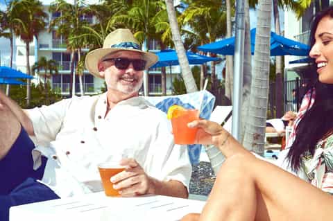 Man in white shirt and straw hat holding orange cocktail while chatting with woman at beachside resort under blue umbrellas
