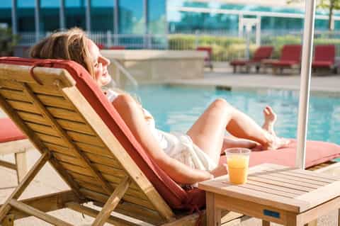 Woman relaxing on wooden lounge chair by resort pool with ocean view and beverage