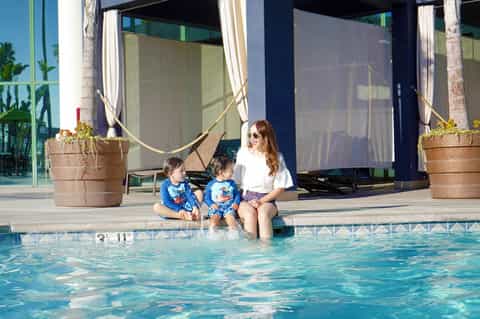 Woman and two children sitting at pool edge with feet in turquoise water, white umbrellas and planters visible