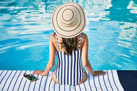 Woman in striped swimsuit and wide-brimmed hat sitting poolside with phone and sunglasses