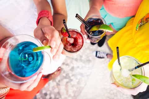 Hands holding colorful tropical cocktails at a beach gathering with umbrellas in background