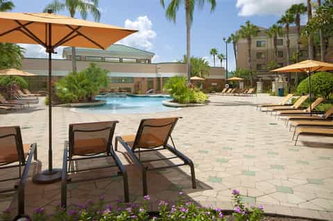 Resort pool area with lounge chairs, orange umbrellas, palm trees, and modern hotel buildings