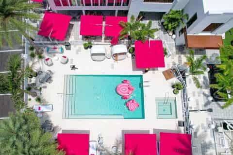 Aerial view of resort pool deck with turquoise water, pink umbrellas, lounge chairs, and tropical landscaping