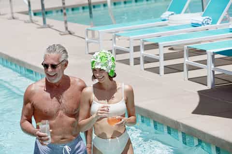 Older couple in swimwear enjoying drinks poolside with lounge chairs and turquoise water visible