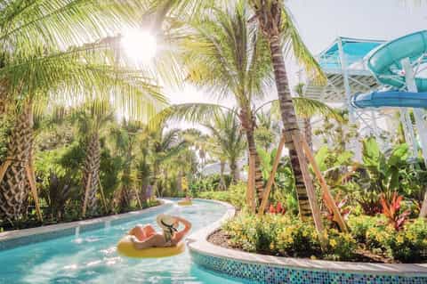 Woman floating on a yellow raft in a lazy river surrounded by palm trees and water slide