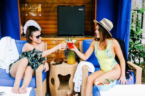 Two women toasting drinks on a blue sofa by a poolside cabana with tropical decor