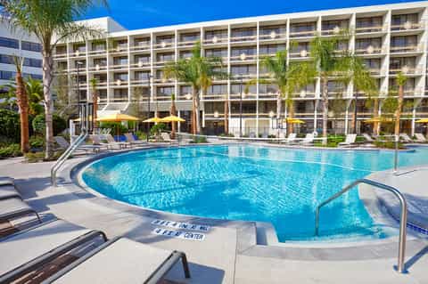 Large resort-style pool with palm trees, yellow umbrellas, lounge chairs, and multi-story hotel building