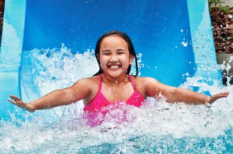 Young girl in pink swimsuit splashing down blue water slide with arms spread