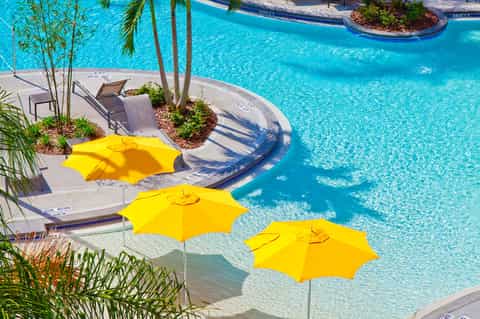 Crystal blue resort pool with yellow umbrellas, lounge chairs, and lush landscaping