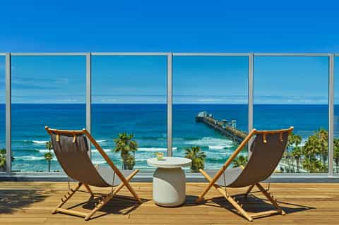 Two beach chairs facing ocean view through floor-to-ceiling windows with pier visible