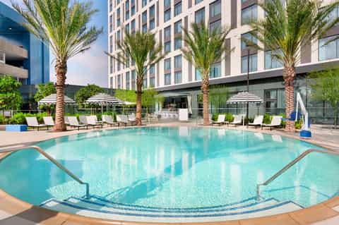 Resort pool surrounded by palm trees, white umbrellas, and modern high-rise hotel building