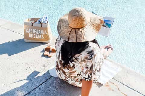 Woman in straw hat sitting poolside with 'California Dreams' beach bag and cocktail
