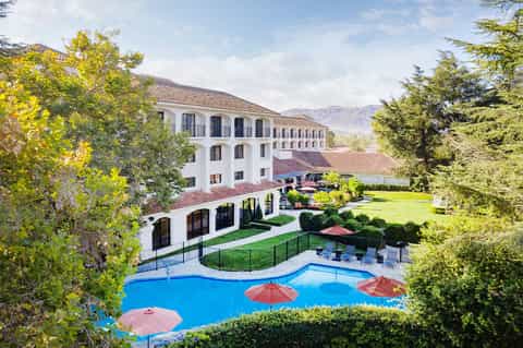 Aerial view of a Spanish-style resort with white buildings, manicured grounds, pool with red umbrellas, and mountain backdrop.