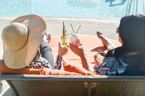 Two women relaxing poolside with cocktails and straw hats overlooking crystal blue water