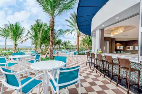 Beachfront patio with turquoise chairs, white tables, palm trees, and ocean horizon view