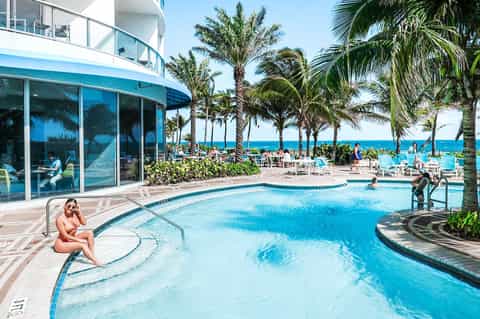 Resort pool overlooking the ocean with beachgoers and palm trees under clear blue sky
