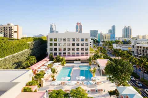 Aerial view of resort pool with pink decking, surrounding palm trees, and city skyline backdrop
