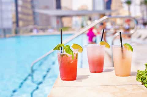 Four colorful tropical cocktails with straws and citrus garnish lined up poolside
