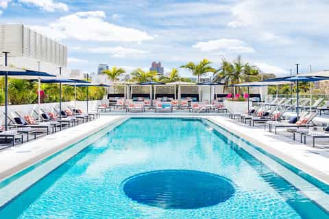 Resort swimming pool with blue water, loungers, umbrellas, and palm trees against city skyline
