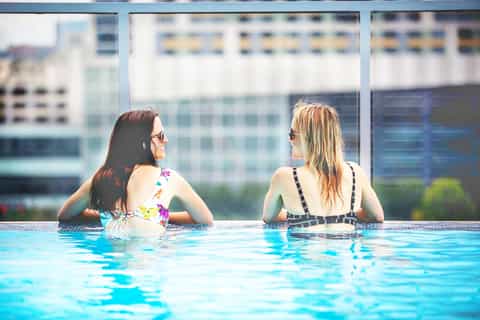 Two women in a rooftop infinity pool overlooking a city skyline with modern buildings