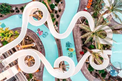 Aerial view of white water slide and turquoise pool with lounge areas and palm trees