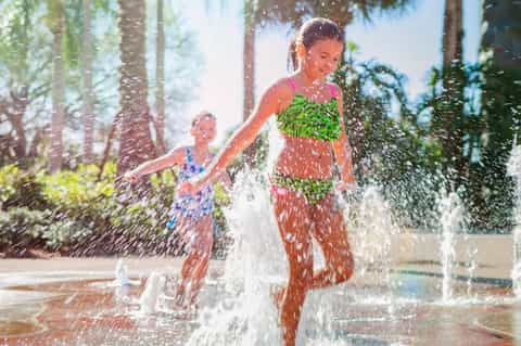 Children playing in outdoor water splash pad surrounded by palm trees on sunny day
