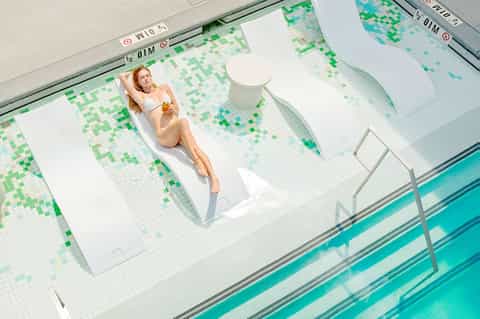 Woman sunbathing on white lounger beside indoor swimming pool with green mosaic tiles
