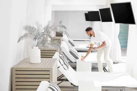 Modern spa lounge with white loungers, potted plants, and staff member serving beverages