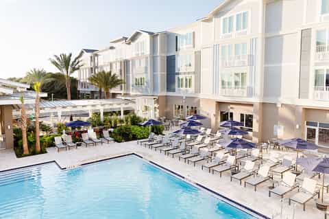 Modern resort pool area with multiple white umbrellas, lounge chairs, palm trees, and contemporary building