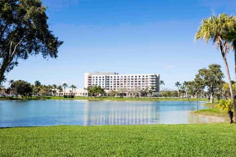 Waterfront hotel building with palm trees, manicured lawn, and calm blue water reflection under clear sky