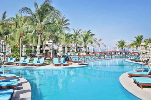 Resort swimming pool with blue water, palm trees, loungers, and ocean view in background