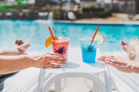 Two colorful tropical cocktails with fruit garnishes on poolside table with swimmers in background