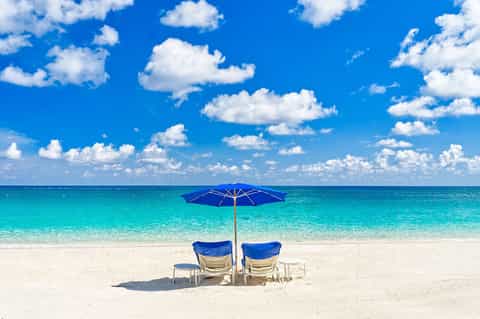 Two blue beach chairs with umbrella on pristine white sand facing crystal turquoise ocean and blue sky