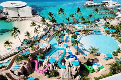 Aerial view of large tropical resort with multiple pools, water slides, blue umbrellas, and turquoise ocean