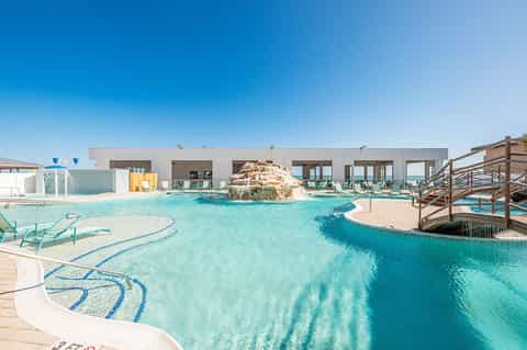 Turquoise resort pool with rock feature, loungers, and modern white building at daytime