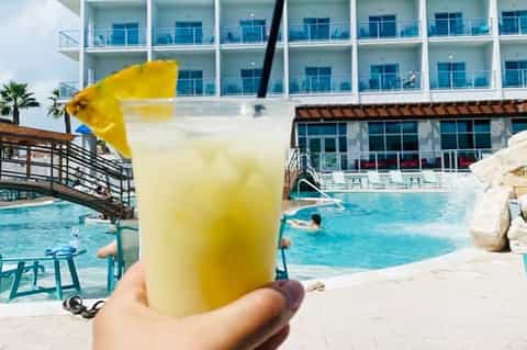 Hand holding yellow tropical drink by resort pool with modern hotel building backdrop