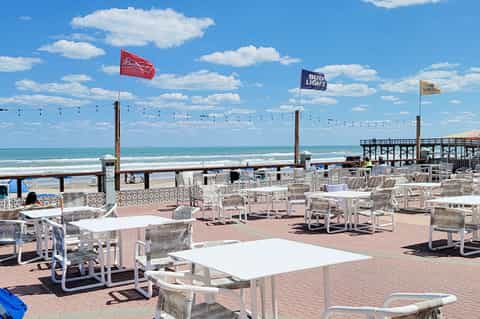 Beachfront dining area with white tables and chairs, ocean view, pier, and blue flag umbrellas