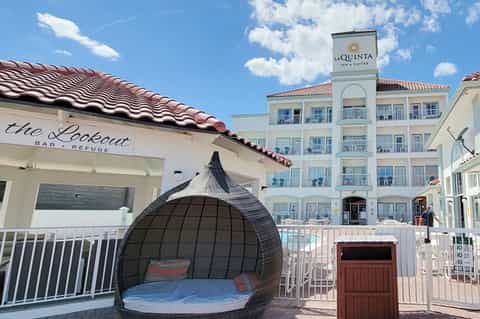 Beach bar lounge with dark pod seating, white railings, and hotel building behind