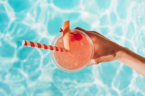 Tropical cocktail with coral-colored drink and striped straw held above sparkling swimming pool water